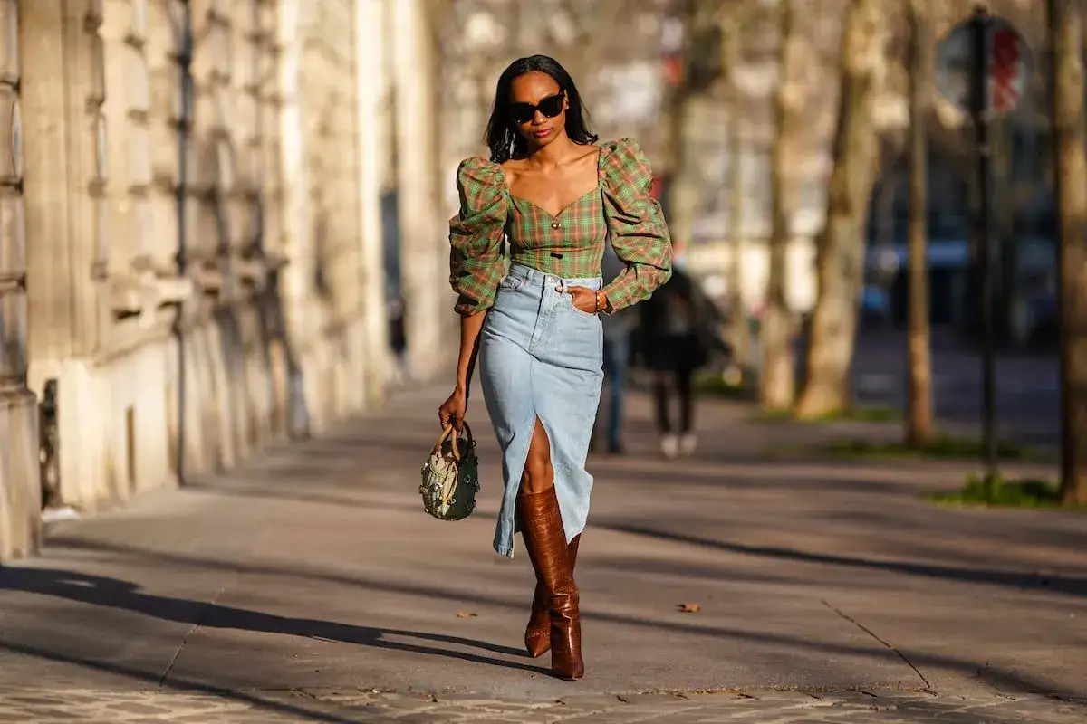 Mujer pasea con una falda vaquera larga, top de cuadros y botas altas.