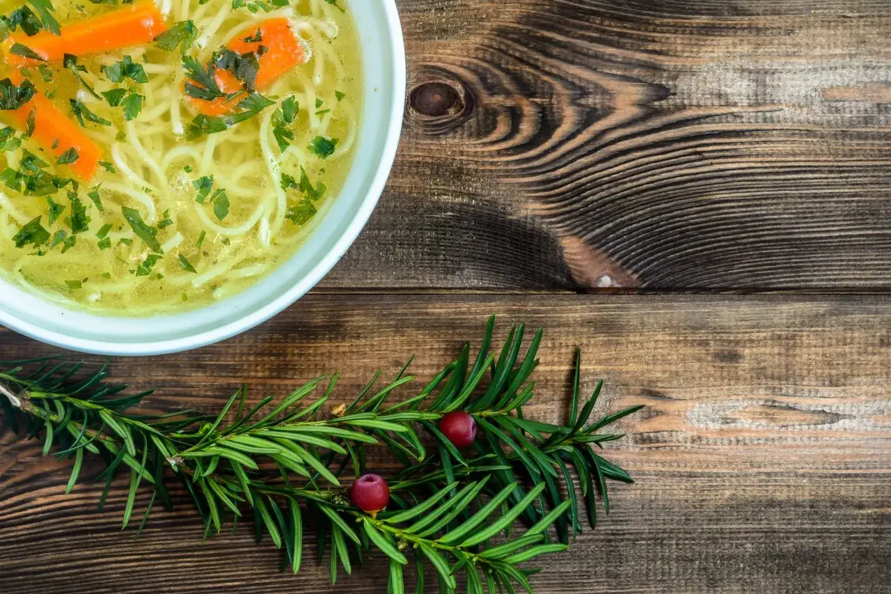 Aromaticzny rosół z makaronem do rosołu, marchewką i natką pietruszki, podany na drewnianym blacie z gałązką iglaka.