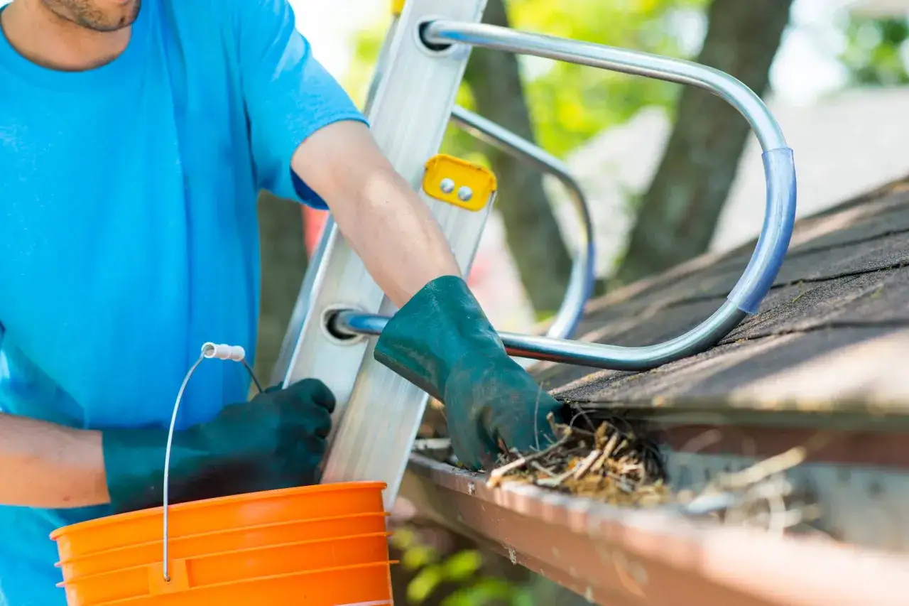 A person in a blue shirt and green gloves cleans debris from a gutter, showing how often should gutters be cleaned to prevent clogs.