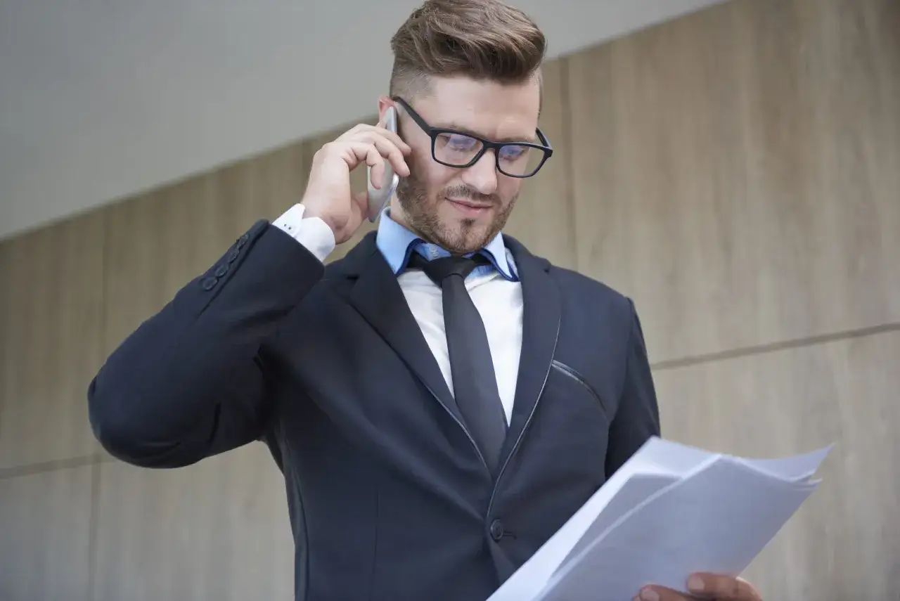 Mężczyzna w garniturze rozmawia przez telefon, trzymając dokumenty. Jego profesjonalny wygląd sugeruje pracę na rynku prawniczym.