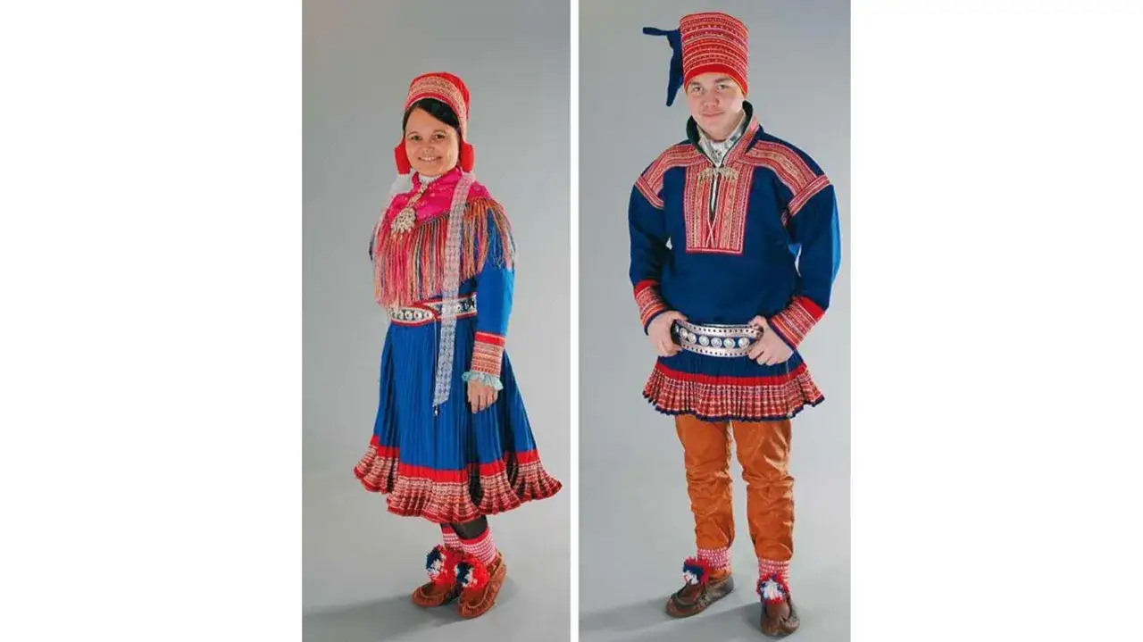 Två personer i traditionella samiska dräkter. Kvinnan bär en blå klänning med röd och rosa utsmyckning, mannen en blå tröja och bruna byxor. Deras samer utseende är färgstarkt och detaljerat.