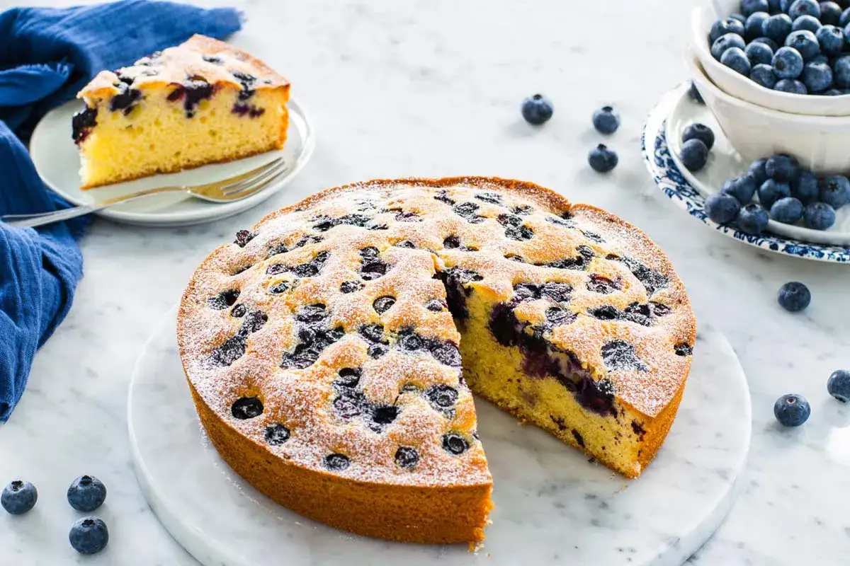 Una deliziosa torta allo yogurt con mirtilli, spolverata di zucchero a velo, su un piano di marmo. Una fetta è già servita.