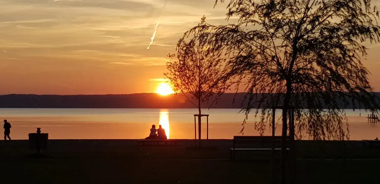Zachód słońca nad największym jeziorem w Austrii. Para siedzi na ławce, podziwiając widok.
