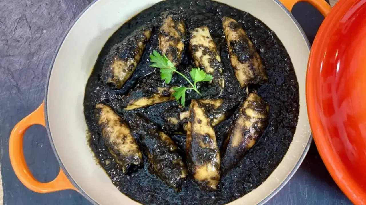 Un plato de calamares en su tinta, con una ramita de perejil, servido en una cazuela de color crema con asas naranjas.