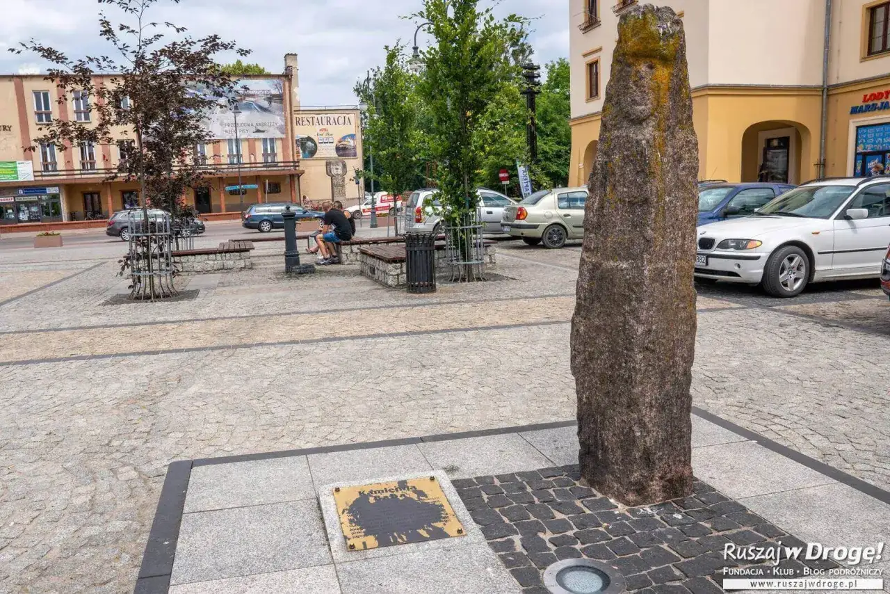 Kamienny obelisk i tablica informacyjna to ciekawe atrakcje na rynku. W tle budynki i zaparkowane samochody.
