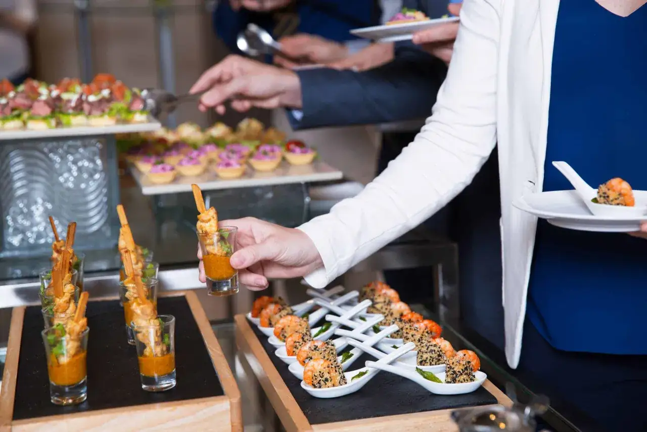 Des convives se servent pour faire un buffet froid. Petites bouchées variées, crevettes et brochettes sont présentées.