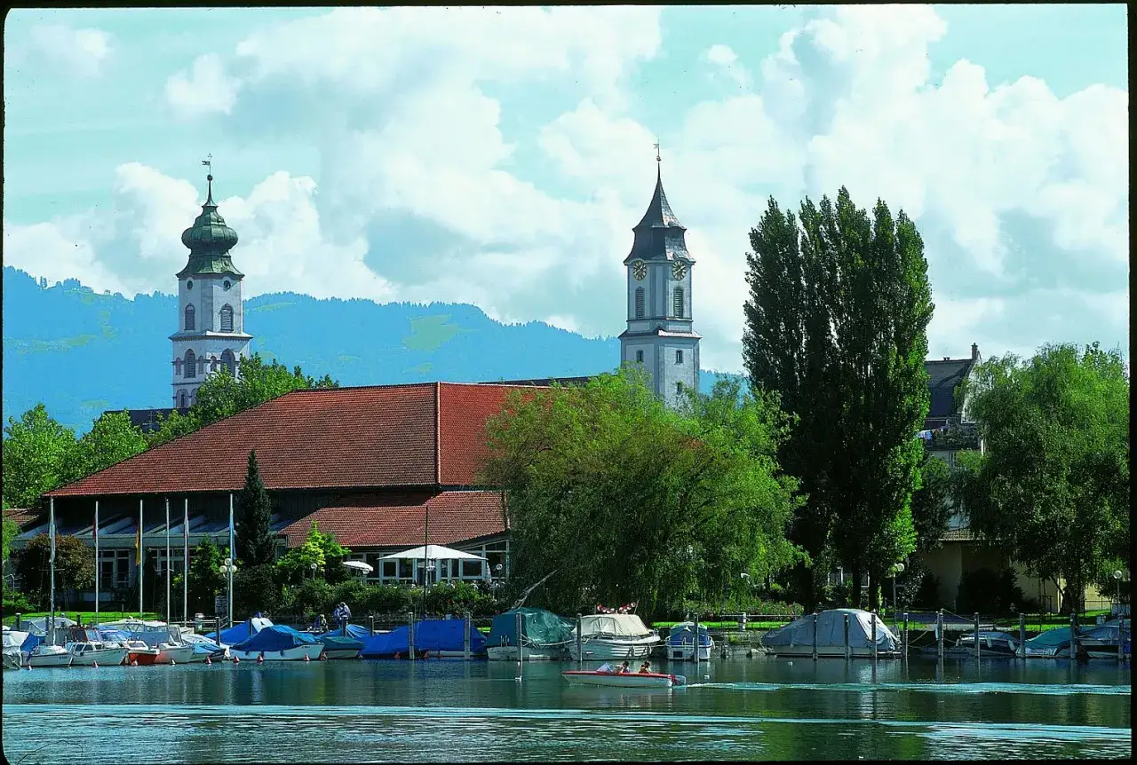 Boote liegen im Hafen vor der Inselhalle Lindau. Im Hintergrund ragen Kircht&uuml;rme und gr&uuml;ne Berge auf.