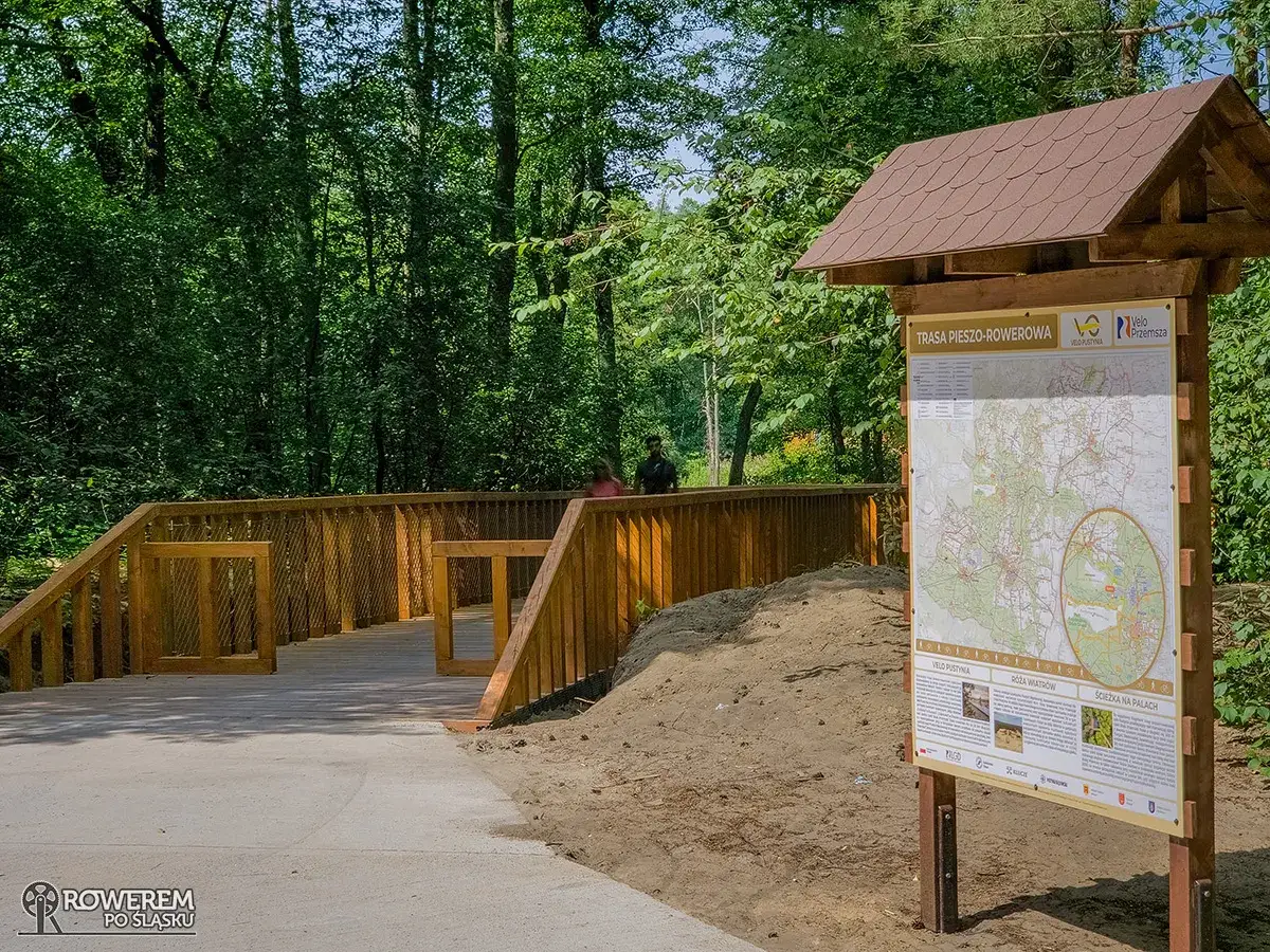 Drewniany mostek i tablica informacyjna z mapą trasy pieszo-rowerowej Velo Przemsza w otoczeniu zieleni.