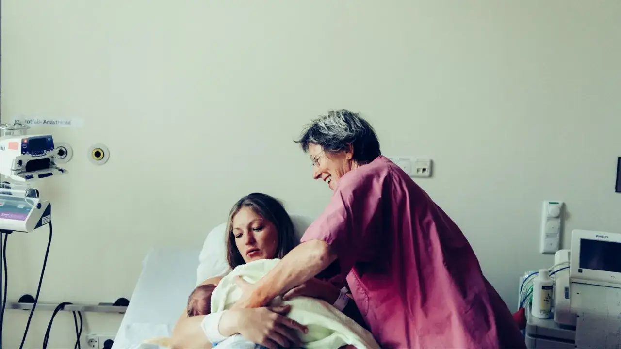 Eine Frau h&auml;lt ihr Neugeborenes, eine Krankenschwester l&auml;chelt. Zeichen, dass die Geburt bald los geht, sind oft subtil, aber die Freude hier ist offensichtlich.