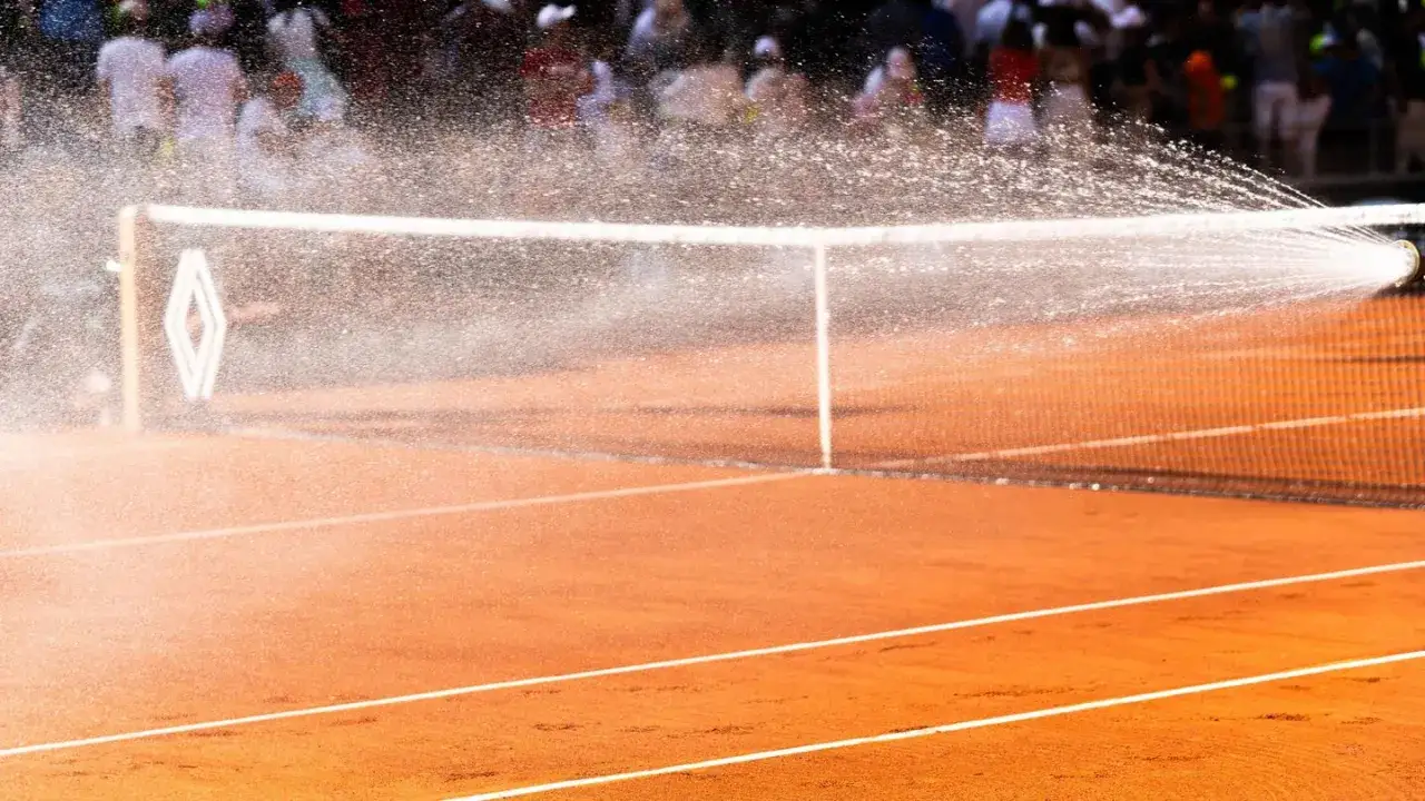 Nawadnianie kortu przed finałem Roland Garros mężczyzn. Widok na ceglaną mączkę i siatkę, w tle rozmyci widzowie.