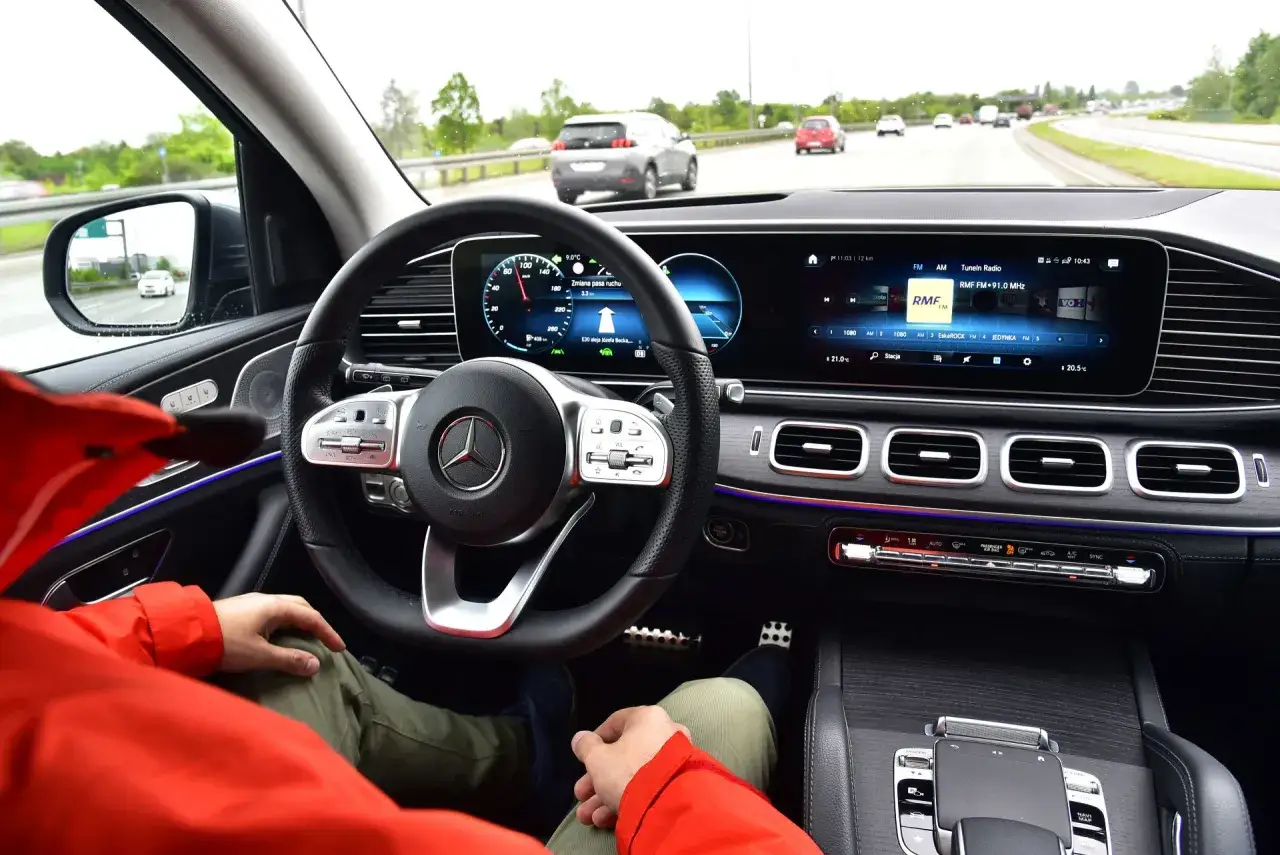 Wnętrze Mercedesa GLE 300d podczas jazdy autostradą. Kierowca w czerwonej kurtce, nowoczesny kokpit z ekranami.