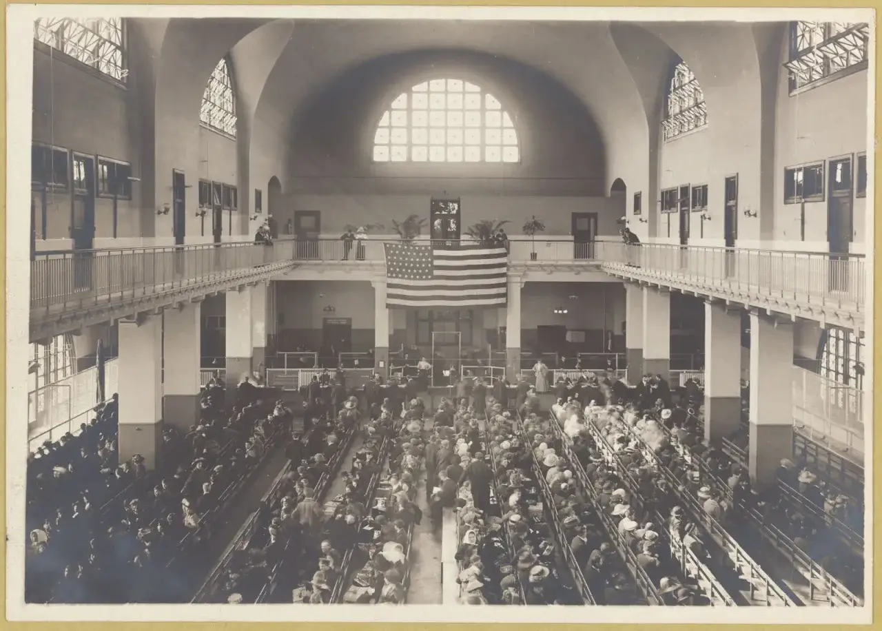 Salle d'attente &agrave; Ellis Island, une foule d'immigrants patiente sous le drapeau am&eacute;ricain.