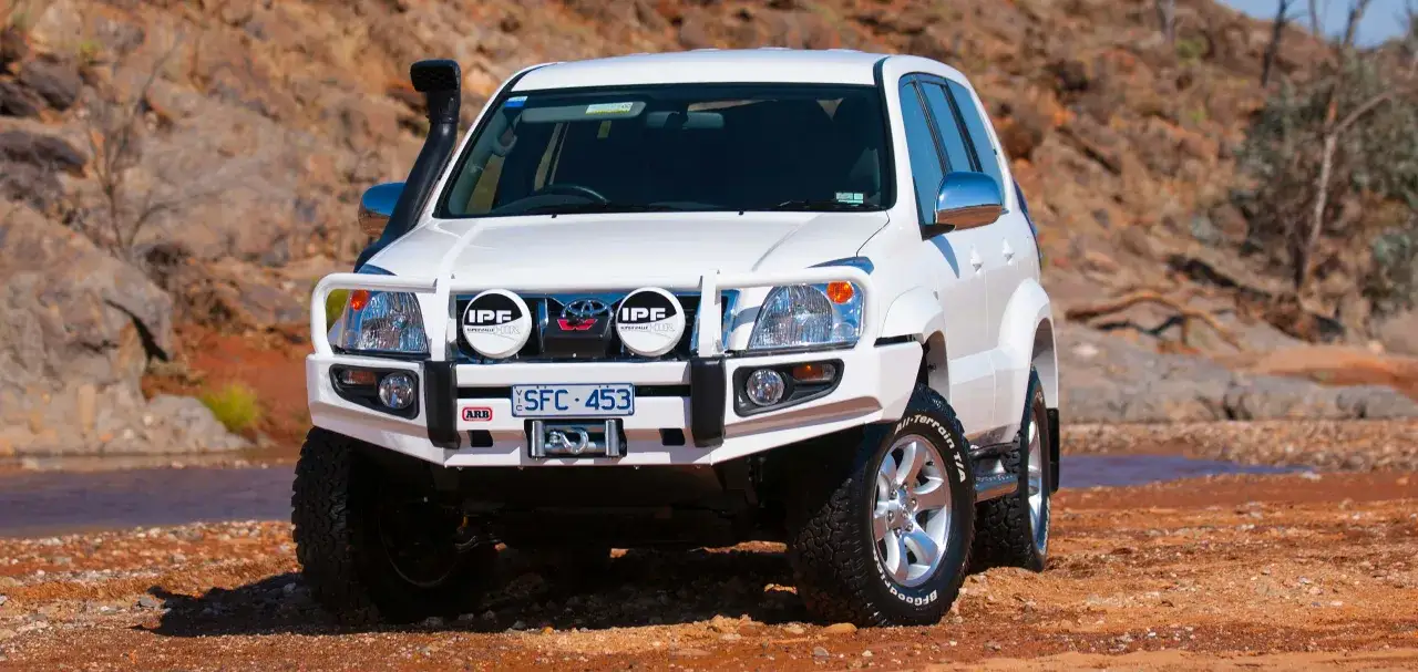 Toyota KDJ 120 blanco, preparado para aventura, con defensa ARB, faros IPF y snorkel, listo para explorar terrenos dif&iacute;ciles.