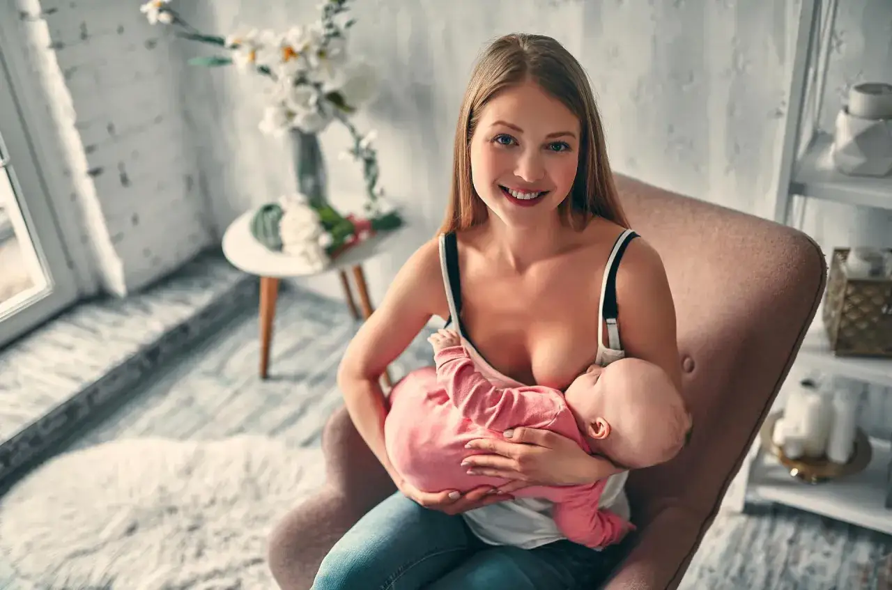 Uśmiechnięta mama siedzi w fotelu, karmiąc piersią swoje dziecko w różowym ubranku.
