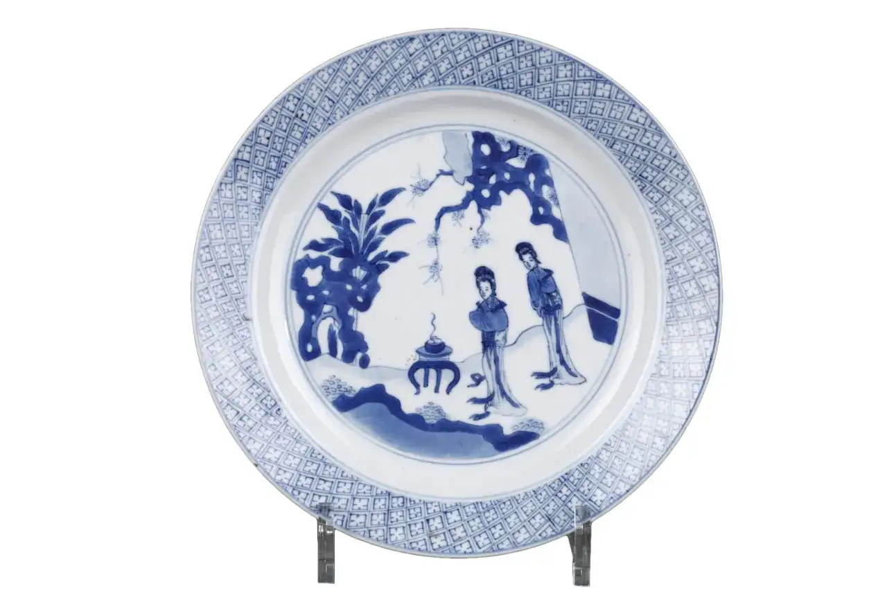 Assiette chinoise ancienne signature, d&eacute;cor&eacute;e de deux femmes dans un paysage stylis&eacute;, bordure orn&eacute;e de motifs g&eacute;om&eacute;triques bleus.