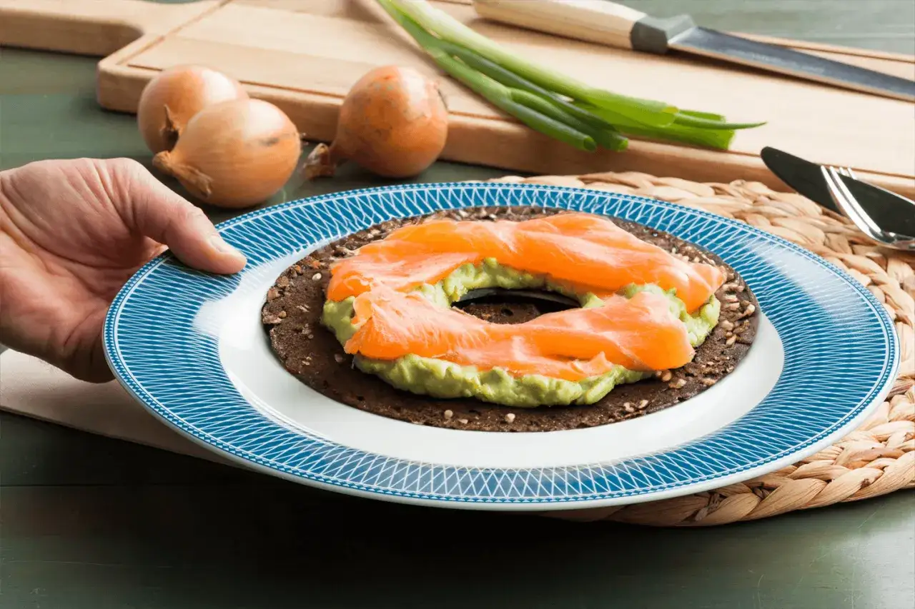 Une main pr&eacute;sente une assiette avec un bagel saumon garni d'avocat cr&eacute;meux et de fines tranches de saumon fum&eacute;.