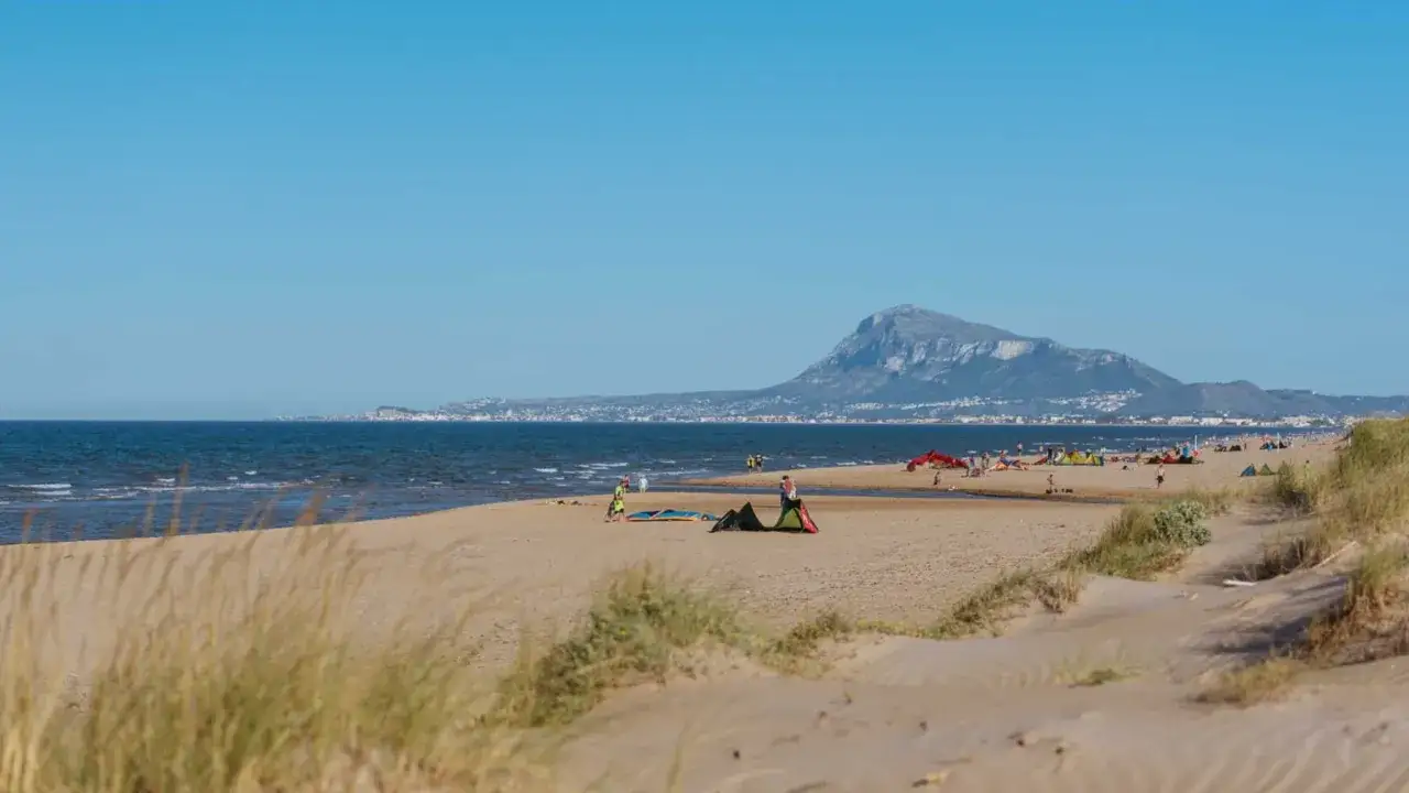 Amplia playa de arena con dunas y vegetación. El mar azul se extiende hasta el horizonte, donde se divisa una ciudad costera y una montaña. Varias personas disfrutan del día, algunas con equipos de kitesurf, en esta **bega de mar**.