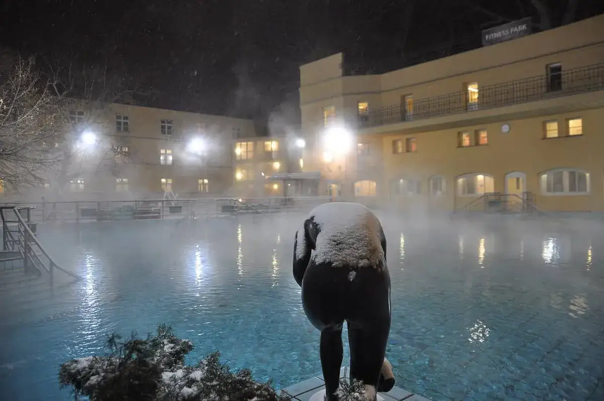Schneebedeckte Statue am Abend vor einem dampfenden Becken im Budapest Thermalbad.