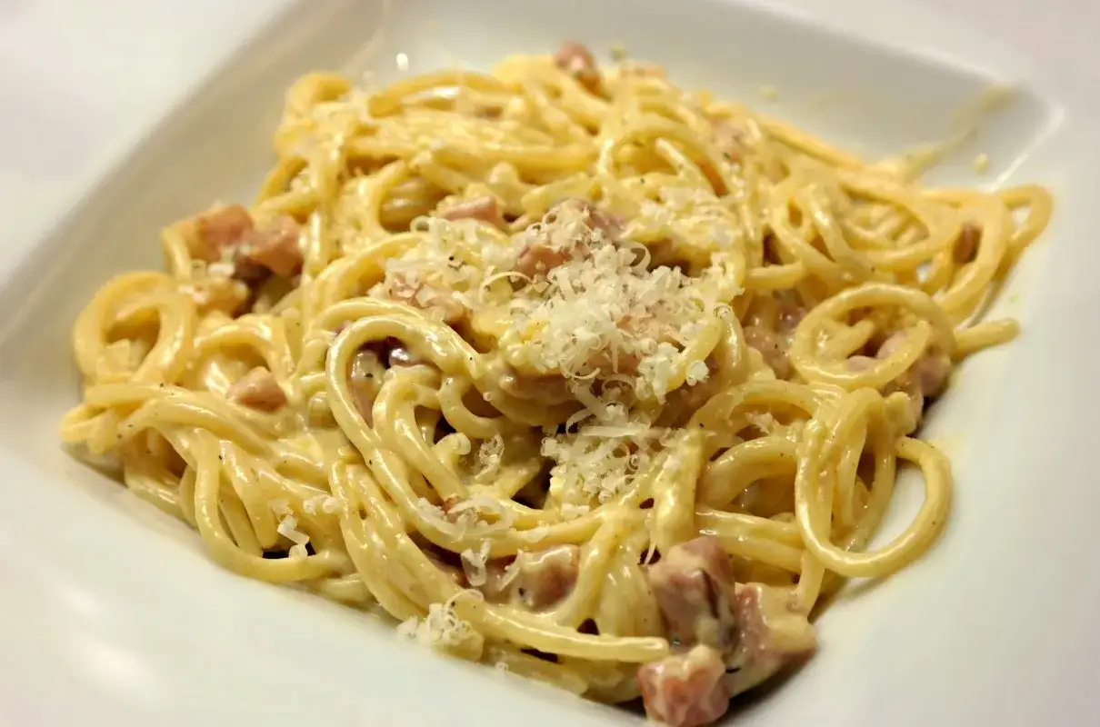 Pyszna carbonara ze śmietaną, podana w białej misce, posypana startym parmezanem i kawałkami boczku.