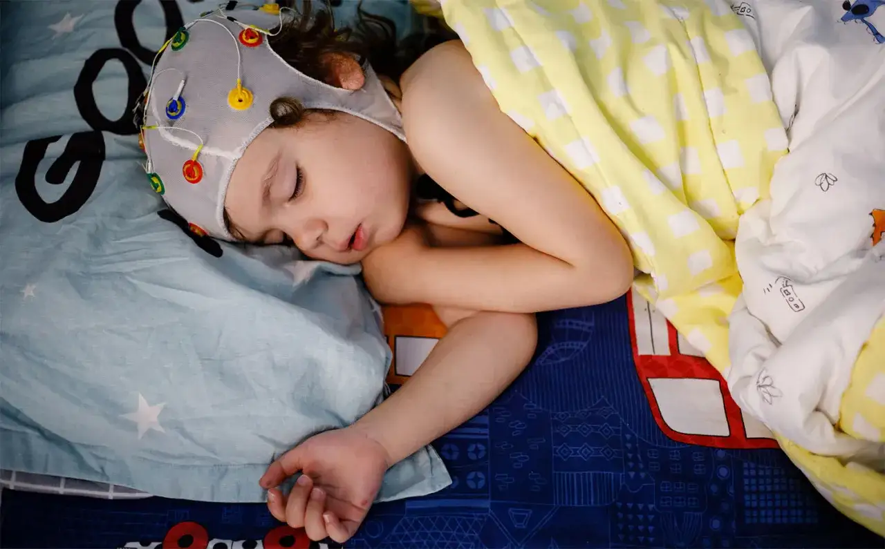 Dziecko śpi w czepku EEG, z kolorowymi elektrodami na głowie. Poduszka i kołdra mają dziecięce wzory.