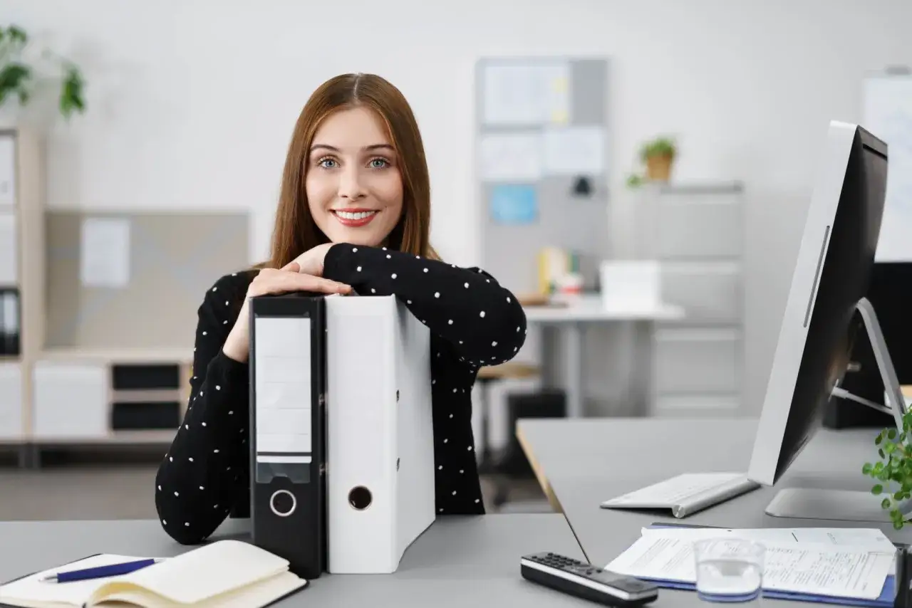 Uśmiechnięta technik administracji opiera się o segregator, gotowa do pracy z dokumentami i komputerem.