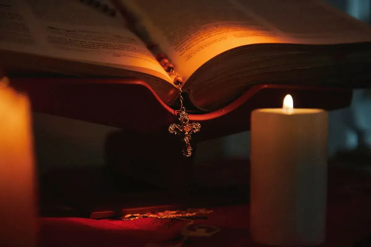 A lit Bible rests on a stand, illuminated by candles. A rosary with a crucifix hangs from the open pages, a symbol of the power in the name of Jesus.