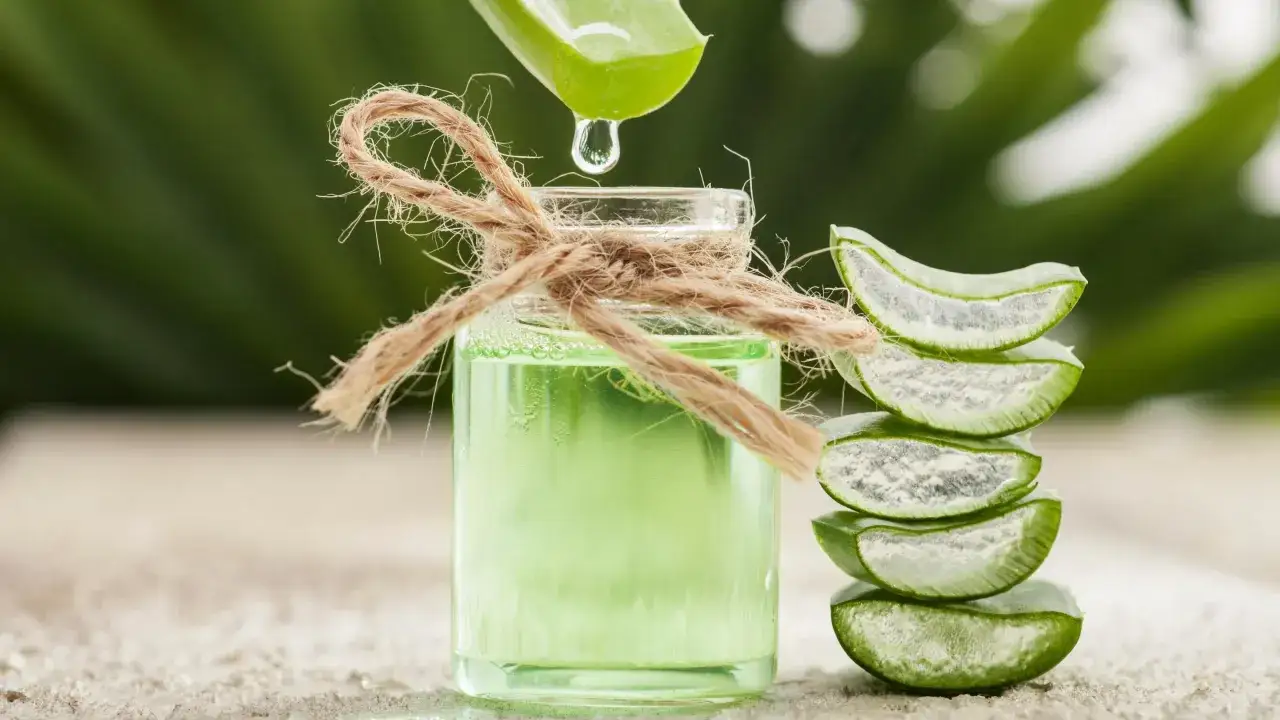 Gotas de aloe vera caen en un frasco con líquido verde, ideal para un suavizante casero para el pelo. Rodajas de aloe apiladas al lado.