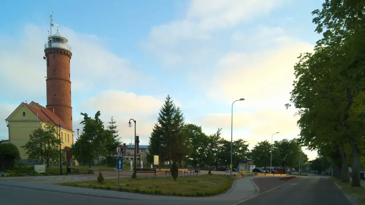 Wysoka, ceglana latarnia morska Jarosławiec g&oacute;ruje nad zielonym placem z ławkami i drzewami.