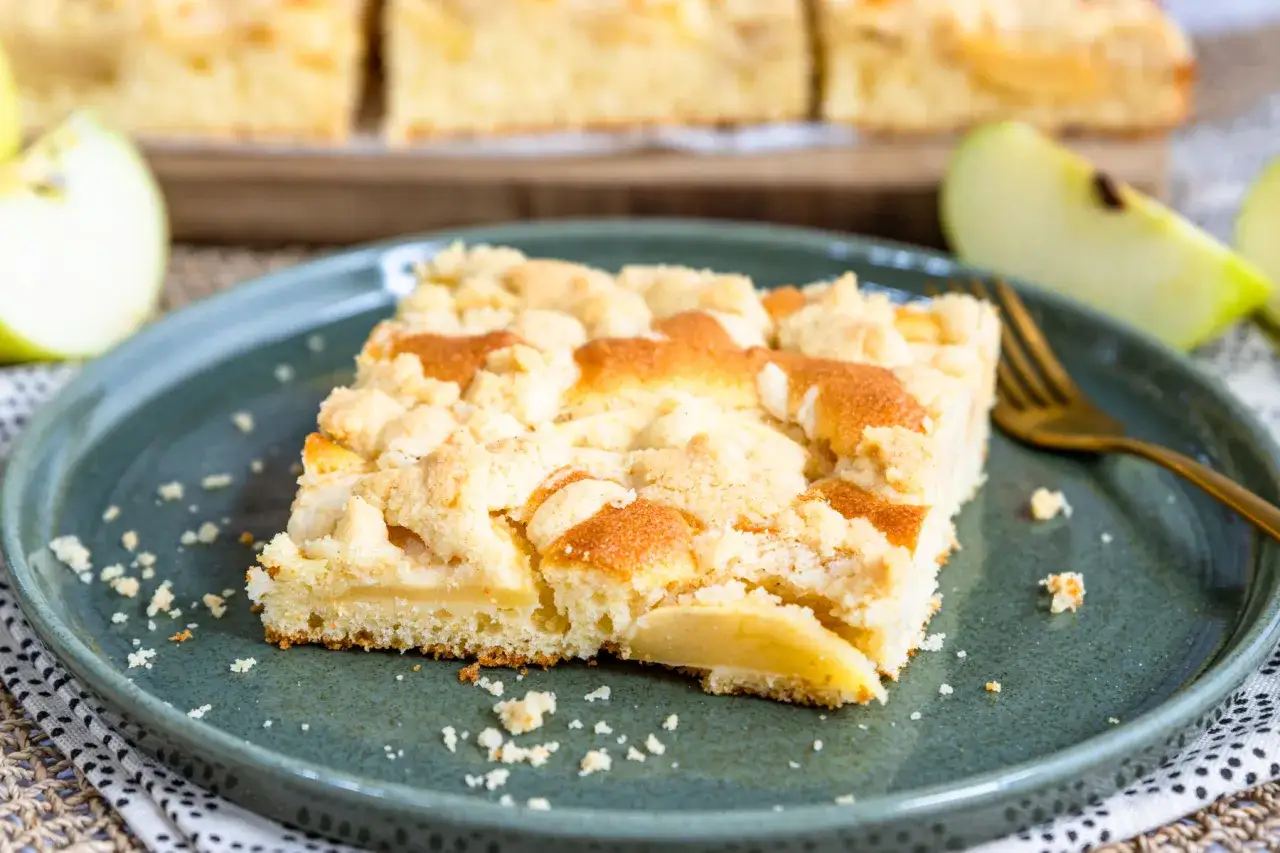 Ein St&uuml;ck vom besten Apfelkuchen mit Streusel, saftig und goldbraun gebacken, liegt auf einem Teller. Frische Apfelspalten im Hintergrund.