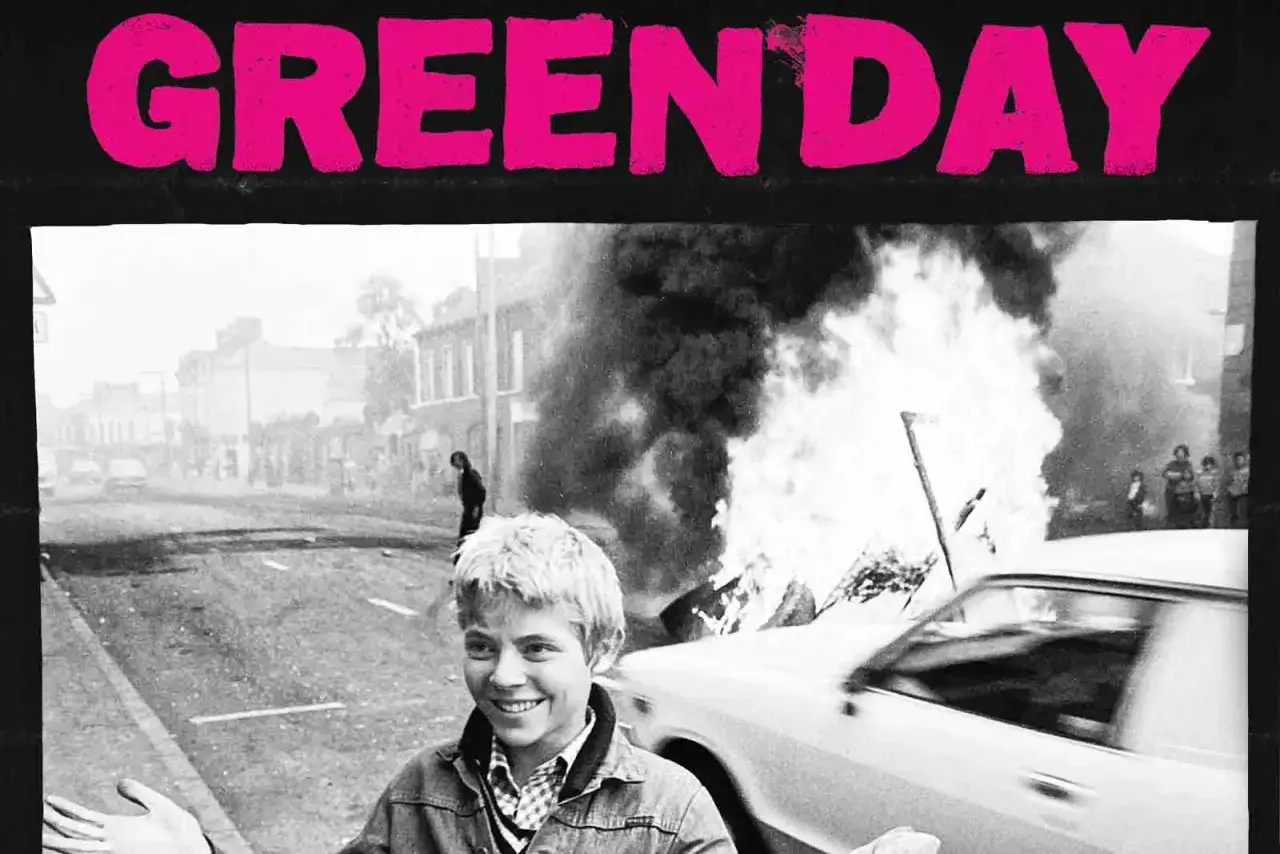 Chłopiec uśmiecha się na tle płonącego samochodu i ulicznych zamieszek, nawiązując do klimatu albumu Green Day Saviors.