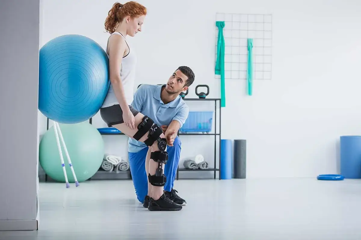 Chinesiologo cosa cura: un uomo assiste una donna con tutore al ginocchio durante un esercizio con palla da fitness.