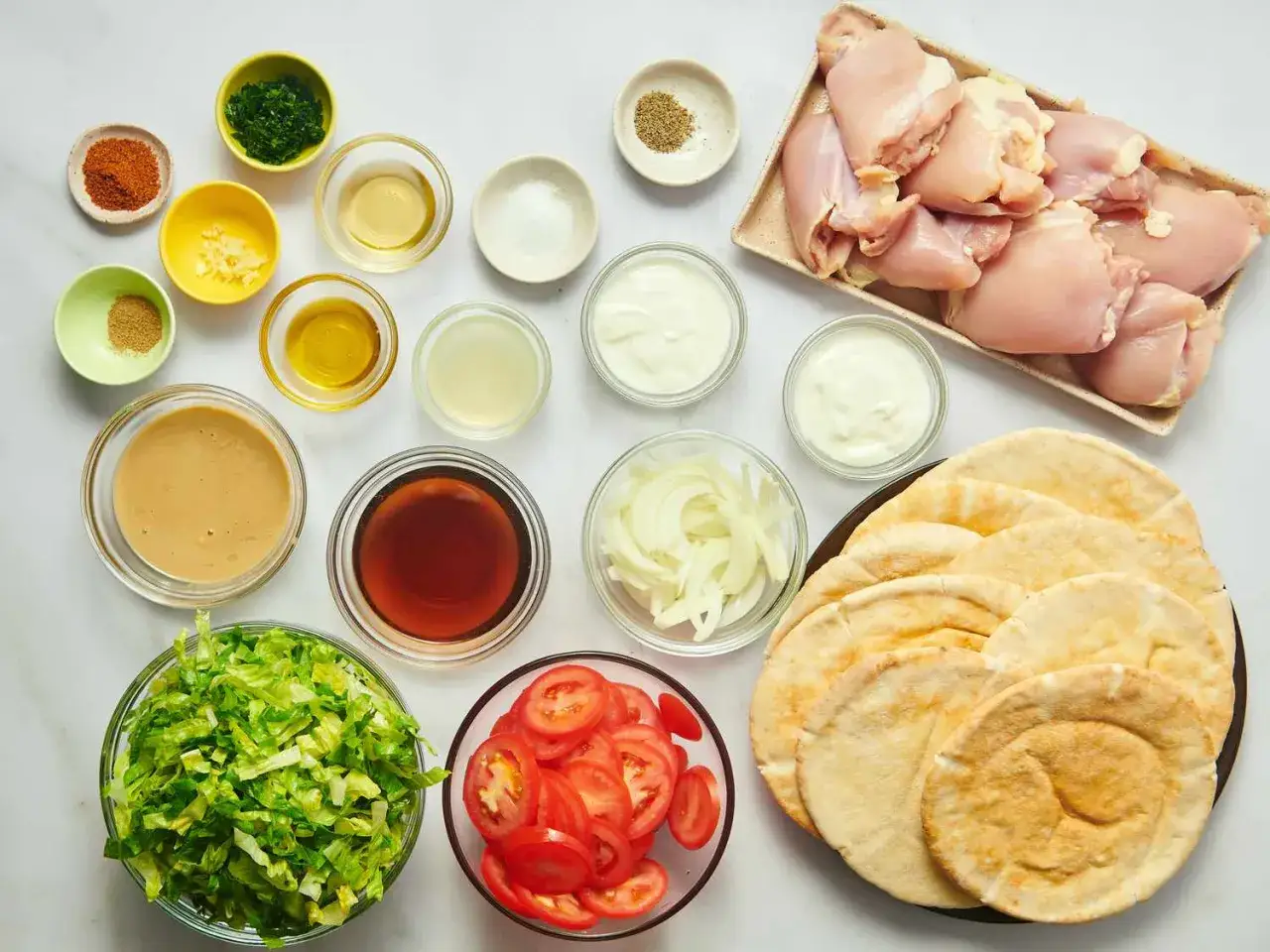 Składniki do przygotowania pysznej shoarmy drobiowej: surowy kurczak, pita, sałata, pomidory, cebula, jogurt, tahini, przyprawy i zioła.