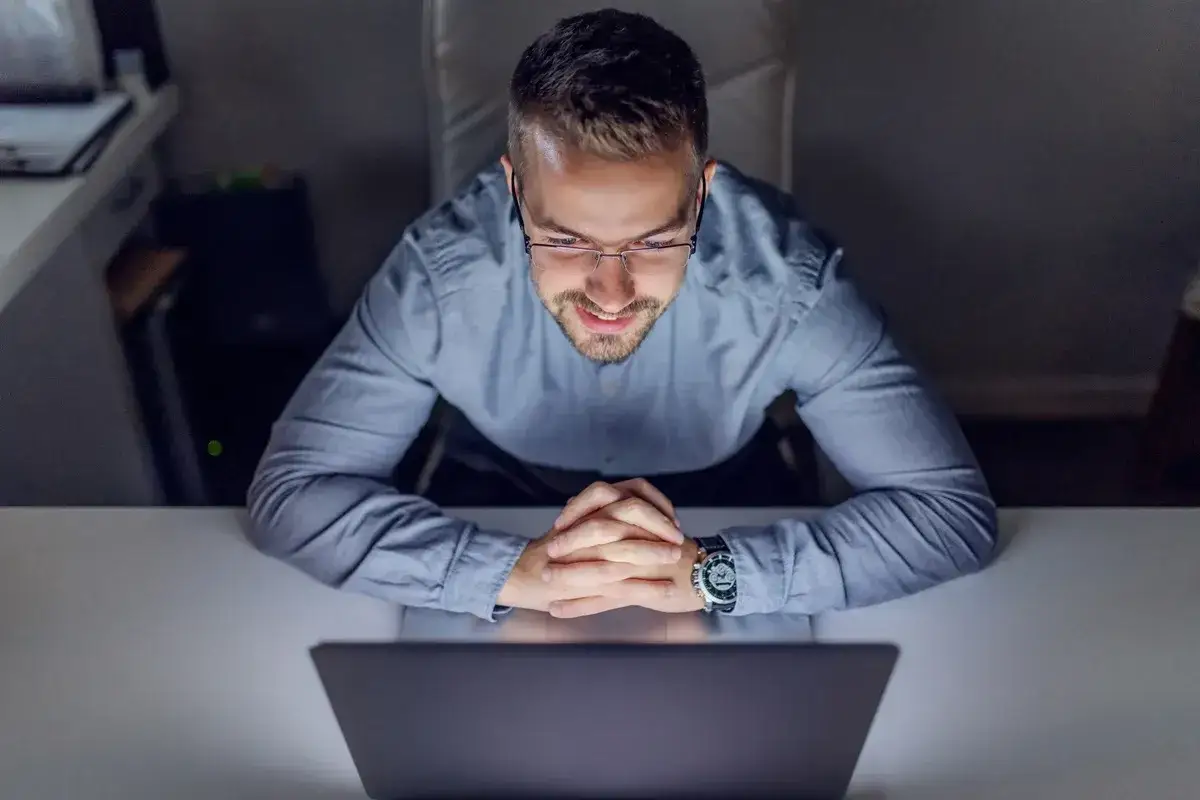 Mężczyzna w okularach pracuje przy laptopie w nocy. Skupiony, z rękami złożonymi na biurku, widać, że jest w trybie zadaniowy czas pracy.