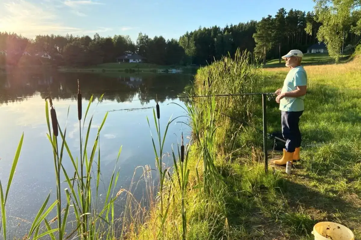 Spokojny wędkarz nad jeziorem Kalwa, czeka na branie ryb. Słońce odbija się w wodzie, a trzciny szumią na brzegu.