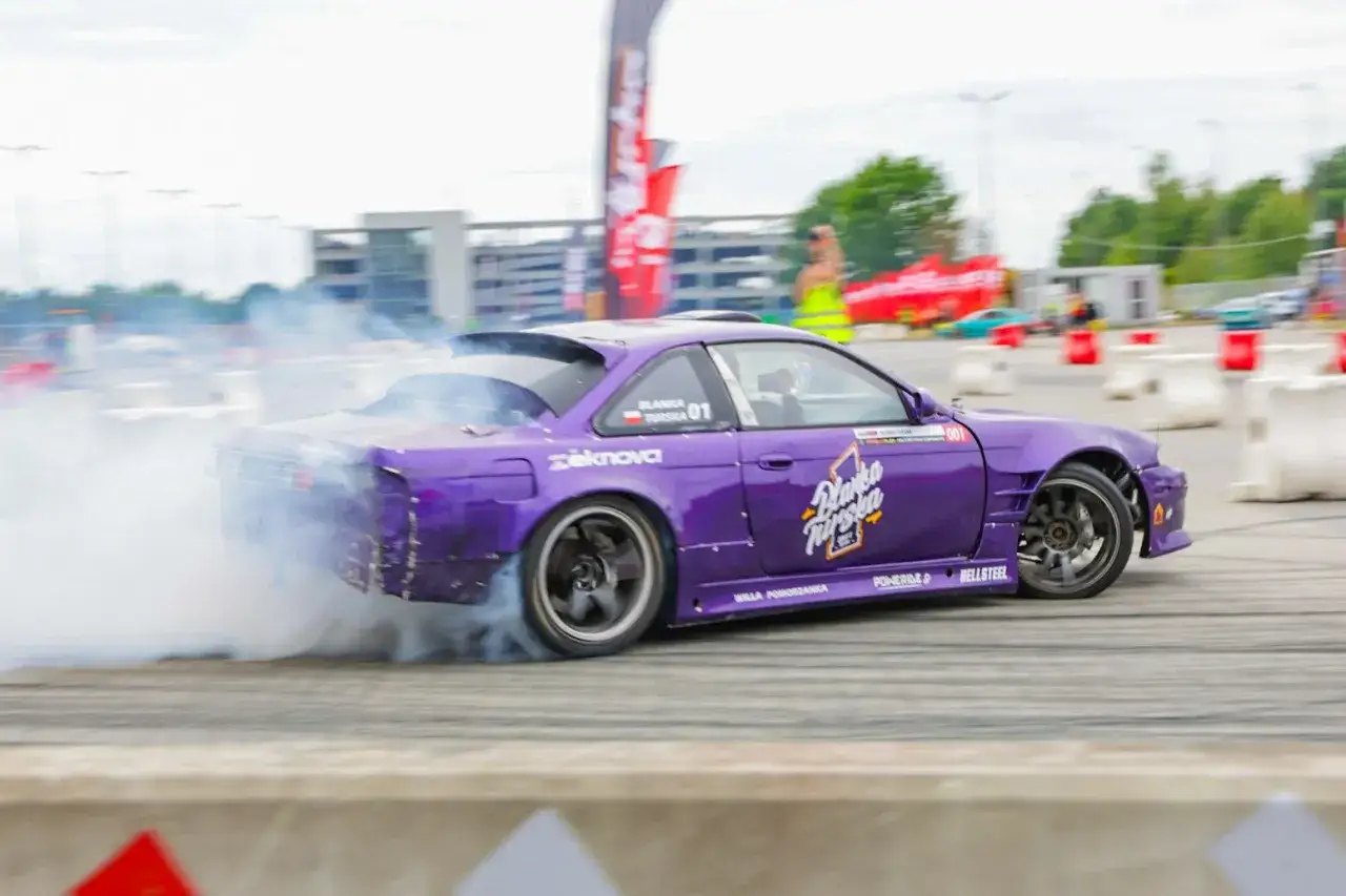 Fioletowe auto do driftu z numerem 01 na drzwiach, w zakręcie z gęstym dymem z opon.