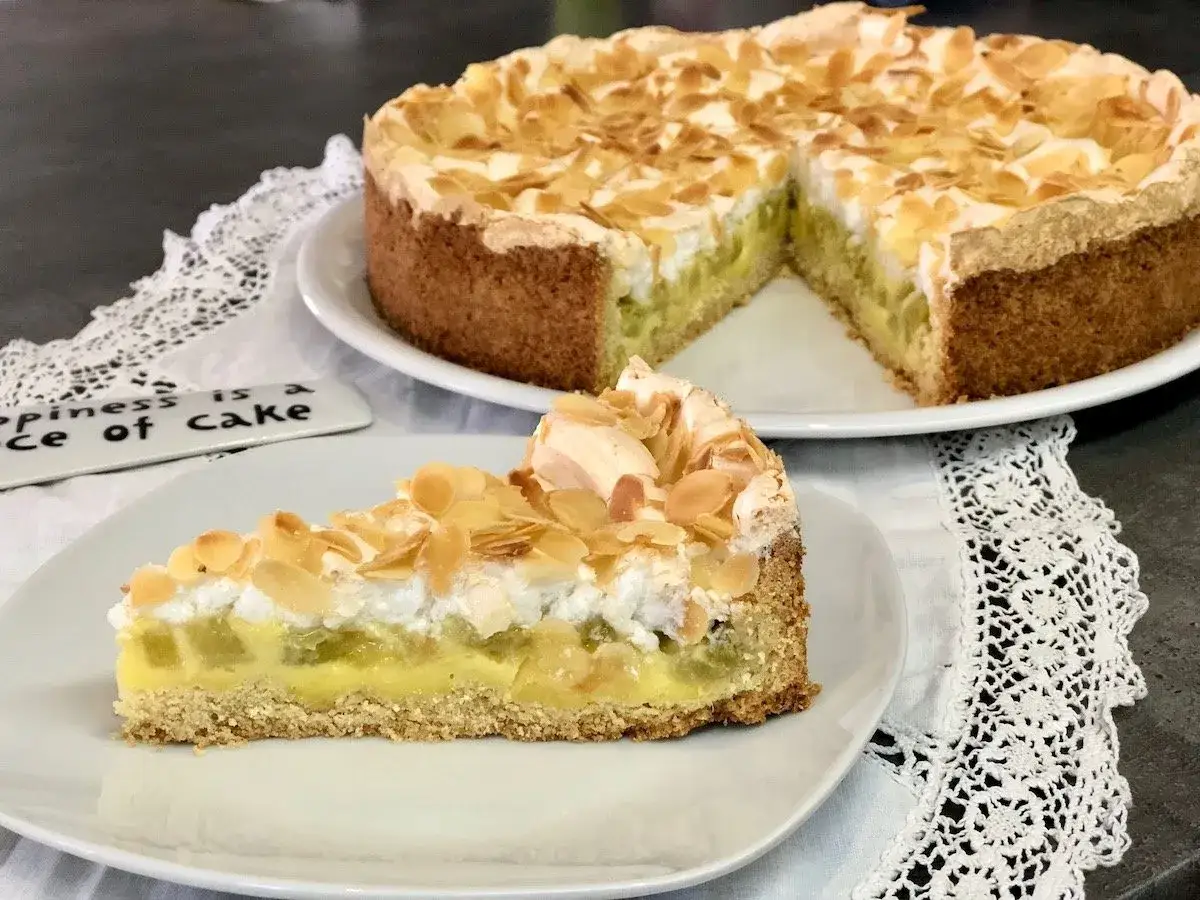 Ein St&uuml;ck Rhabarberkuchen mit Pudding und Baiser, bestreut mit Mandelbl&auml;ttchen. Der ganze Kuchen steht im Hintergrund.