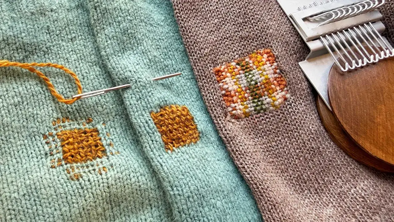 Learning how to darn is a useful skill. This image shows a needle and orange yarn on a teal sweater, with a small woven patch. A brown sock with a colorful patch and a darning mushroom are also visible.