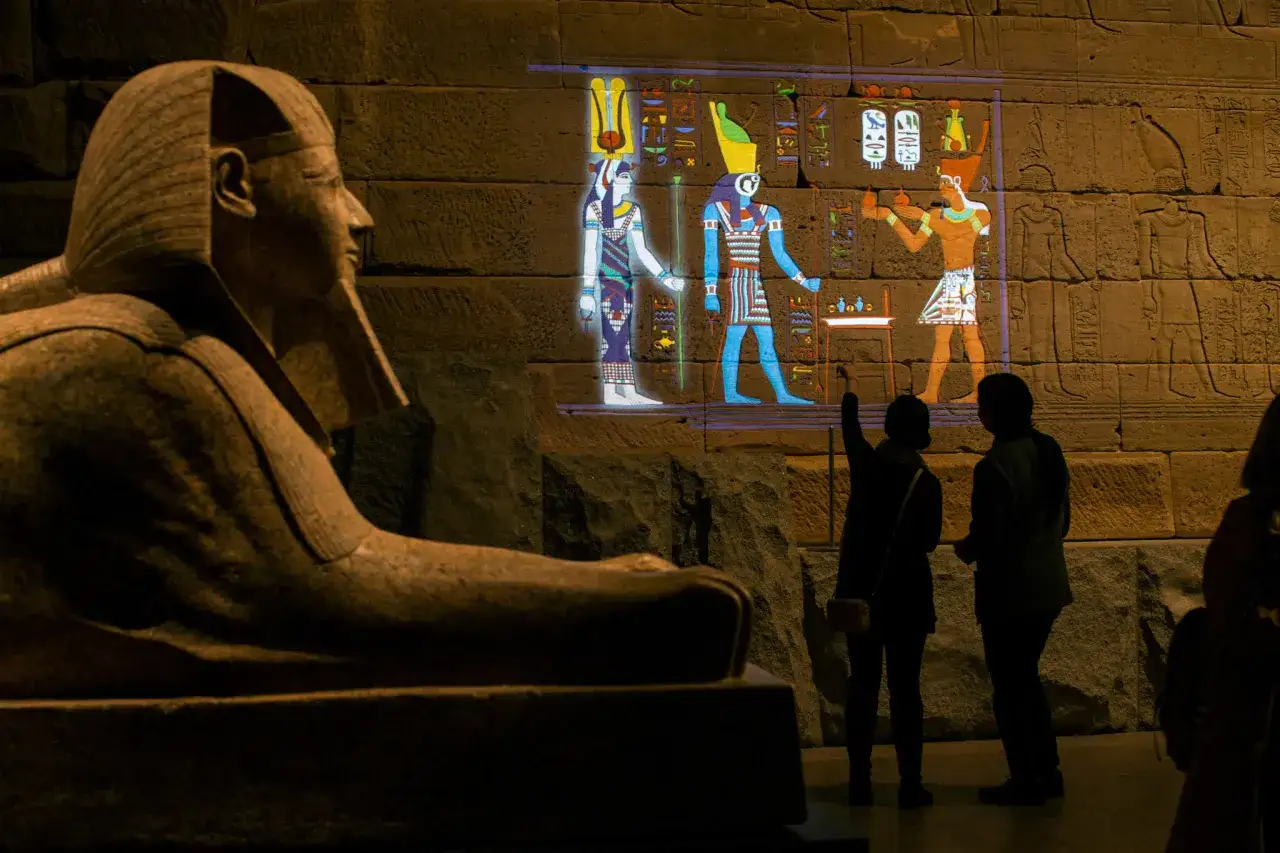 A statue's head and shoulders are in the foreground of the Temple of Dendur. Projections of ancient Egyptian figures illuminate the stone wall behind.