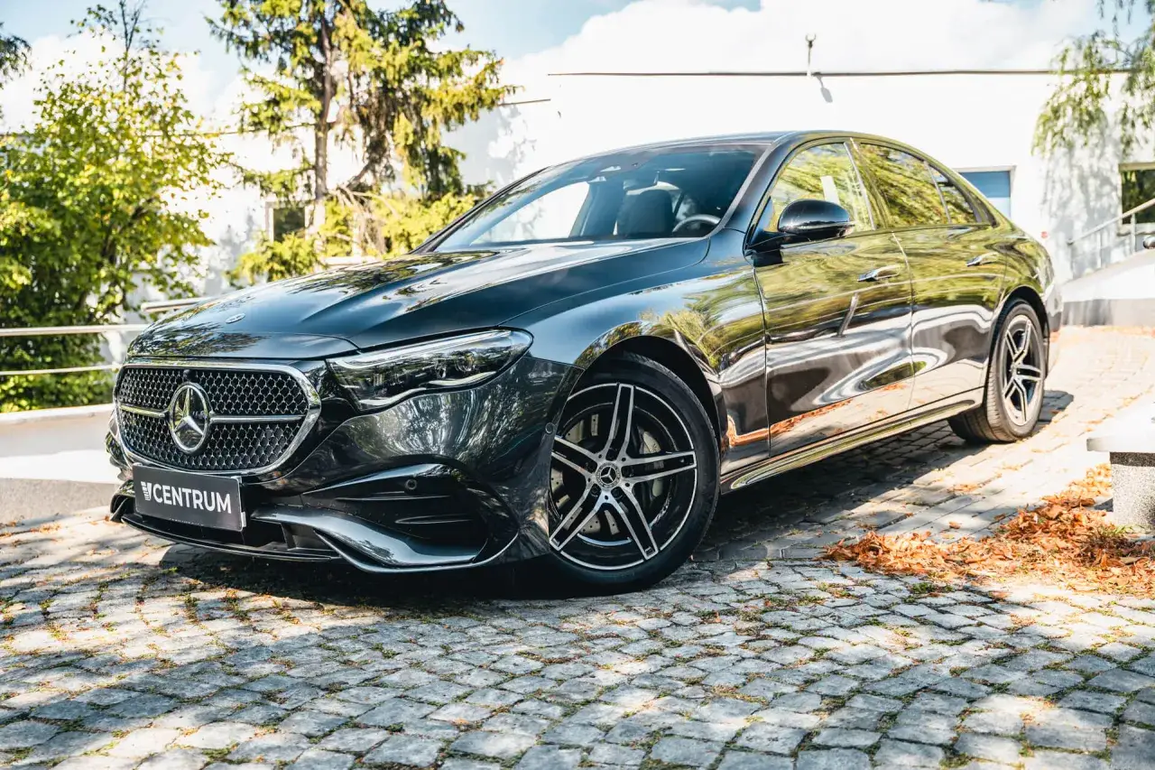 Elegancki, czarny Mercedes 220 błyszczy na brukowanym podjeździe, otoczony zielenią i jesiennymi liśćmi.