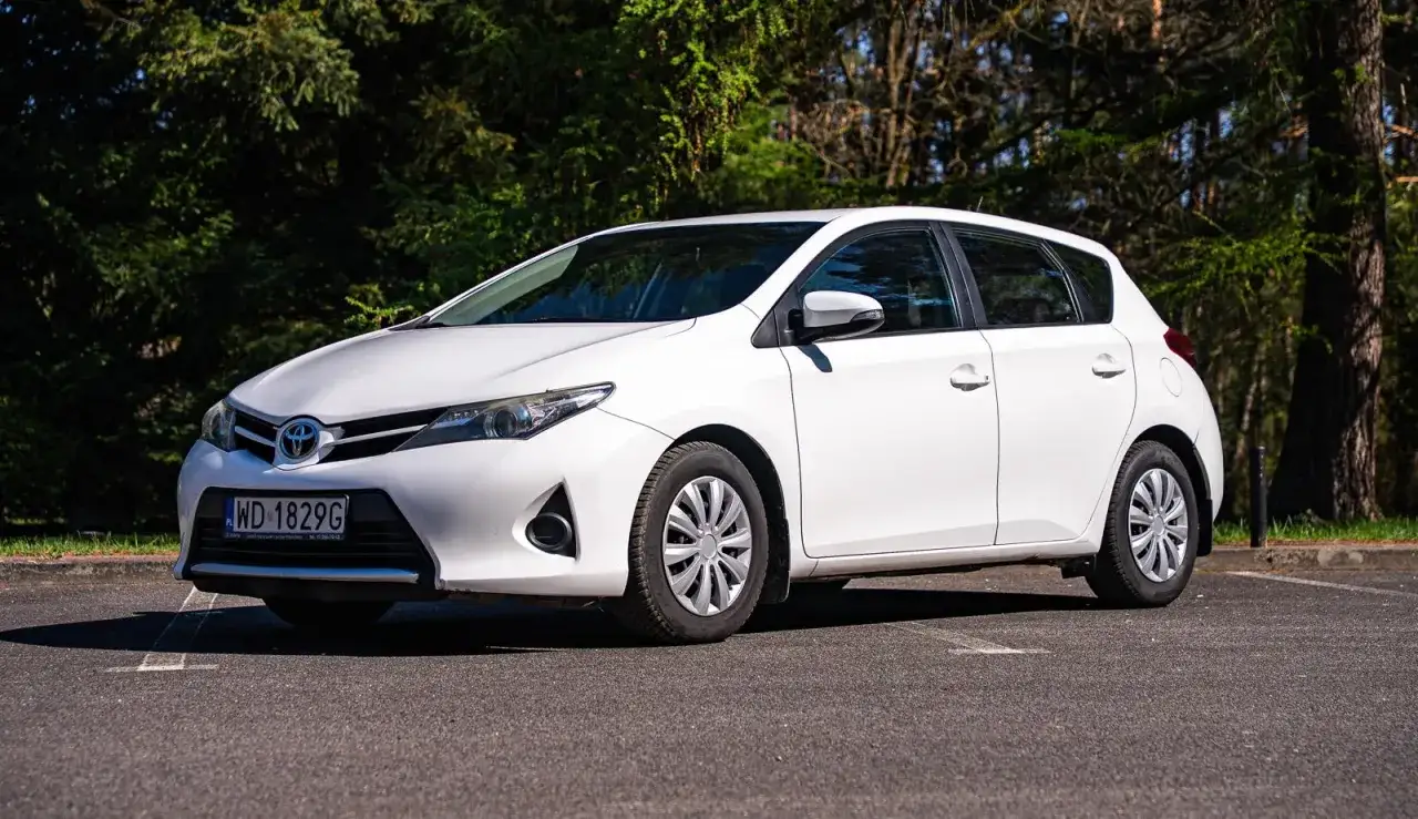 Biały Toyota Auris 2015 zaparkowany na tle drzew.