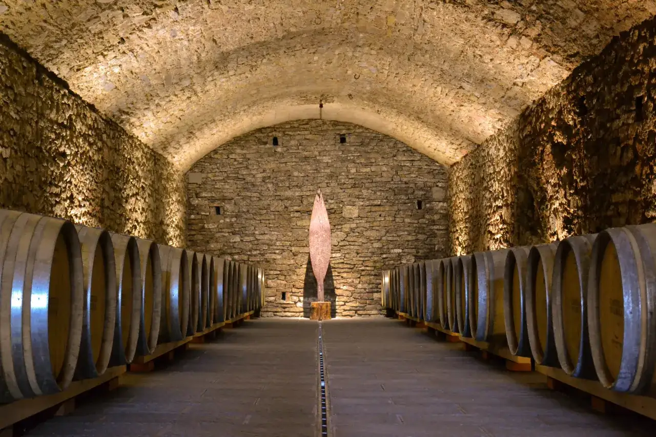 Cantina sotterranea con botti di legno per l'invecchiamento dei vini italiani. Al centro, una scultura moderna.