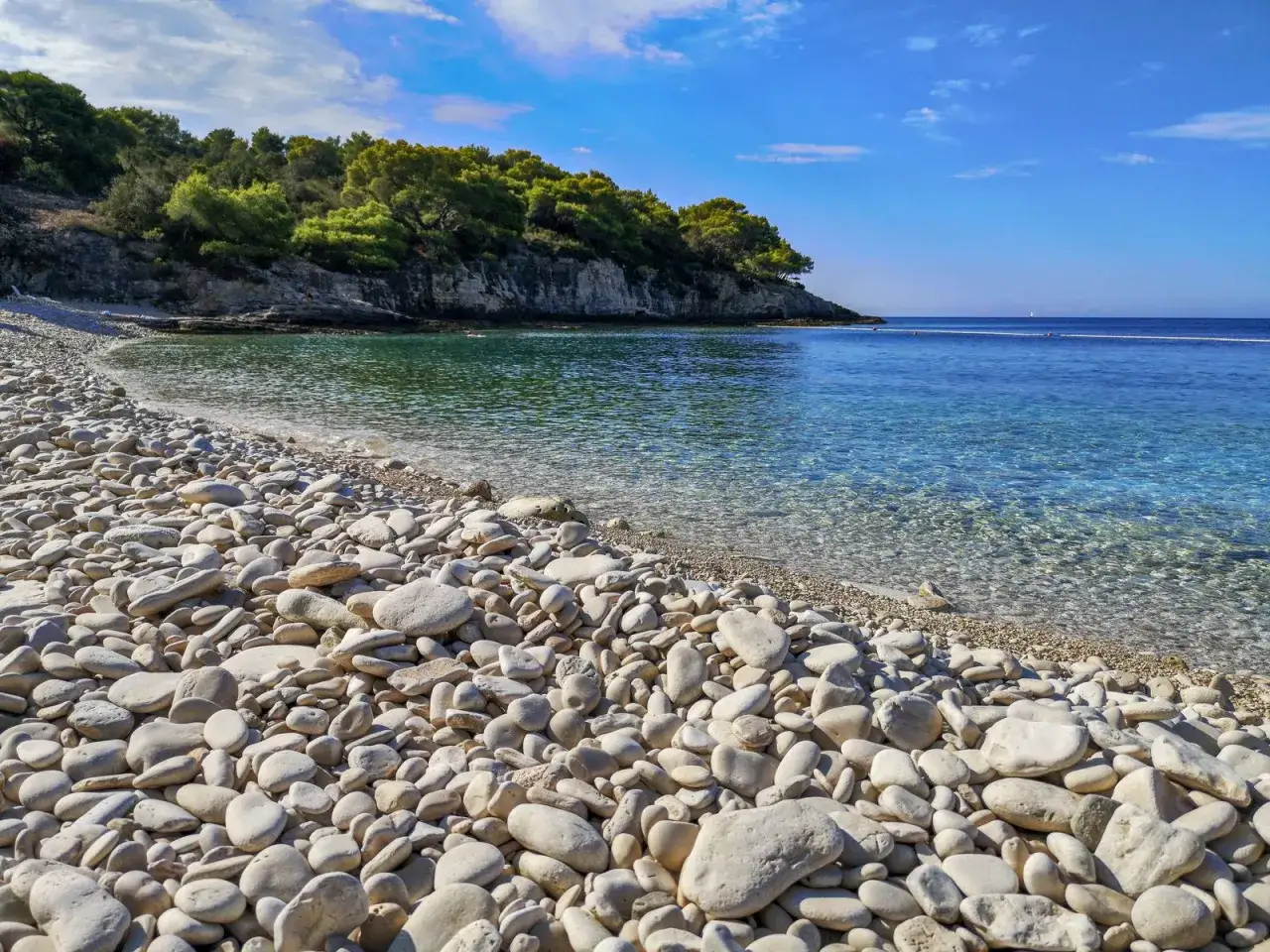 Ein sonniger kroatischer Strand mit Kieselsteinen und klarem, t&uuml;rkisfarbenem Wasser, ges&auml;umt von gr&uuml;nen Pinien.