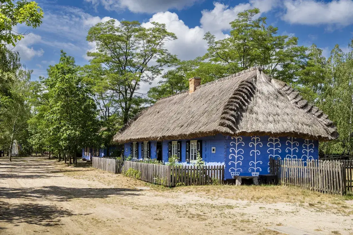 Skansen wsi łowickiej: niebieski dom z białymi wzorami i strzechą, otoczony drewnianym płotem i zielenią.