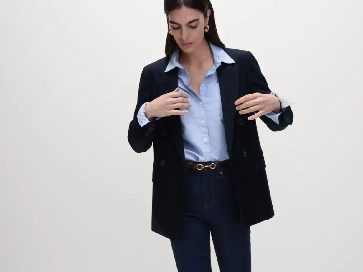 A woman models a business casual look: navy blazer, light blue button-down shirt, dark wash jeans, and a gold bit belt.