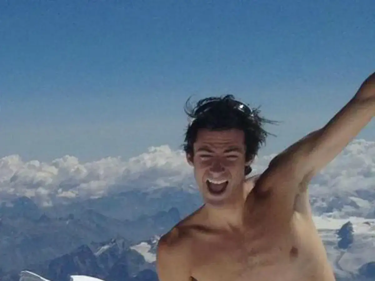 Kilian Jornet, sin camiseta, celebra en la cima del Mont Blanc, con un cielo azul y nubes de fondo.