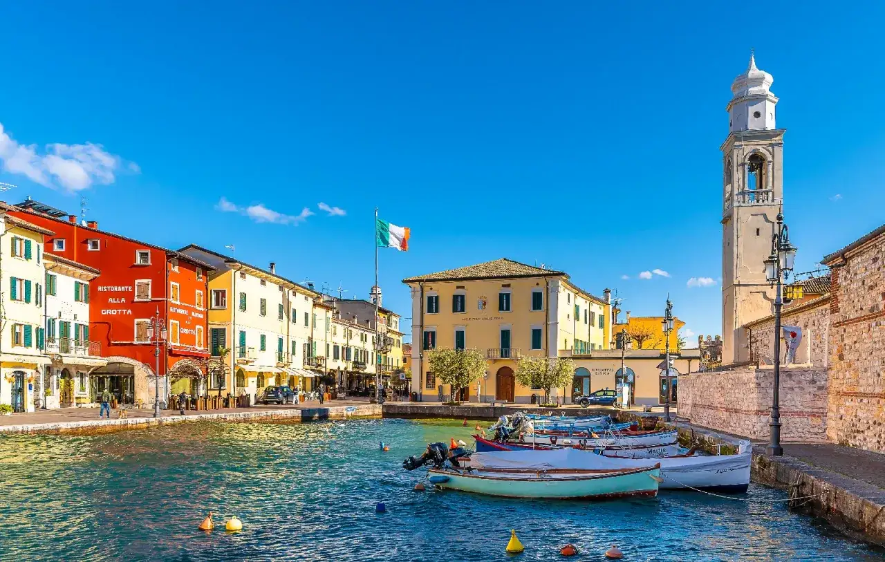 Lazise cosa vedere: un pittoresco porto con case colorate, barche ormeggiate e un'antica torre campanaria sotto un cielo azzurro.