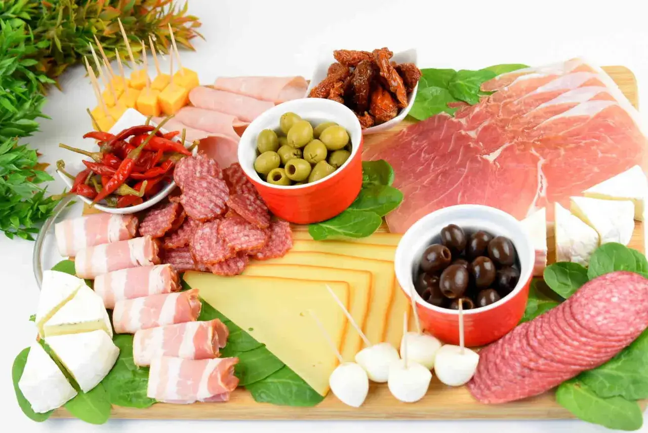 Deska serów i wędlin: szynka crudo, salami, oliwki, suszone pomidory, papryczki chili, ser żółty i biały.