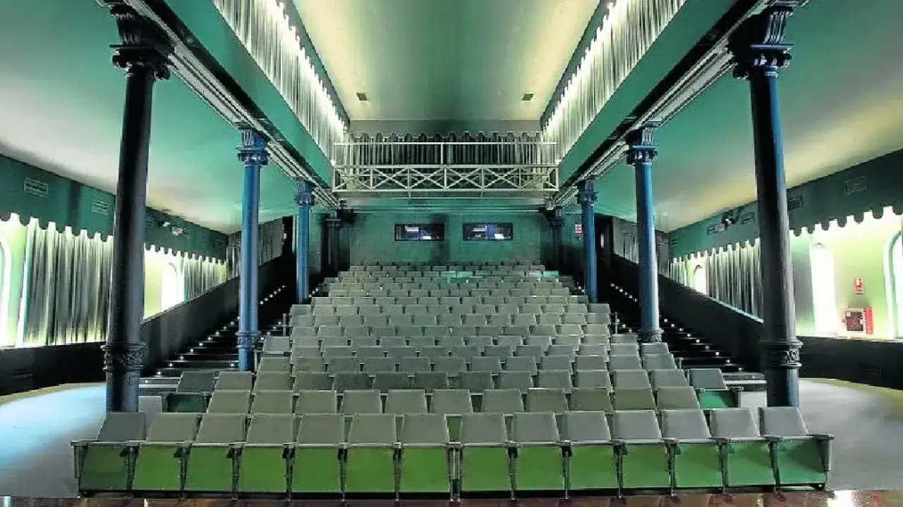 El interior del teatro Mercado de Zaragoza, con sus filas de asientos verdes y grises, columnas oscuras y una barandilla metálica en el nivel superior.
