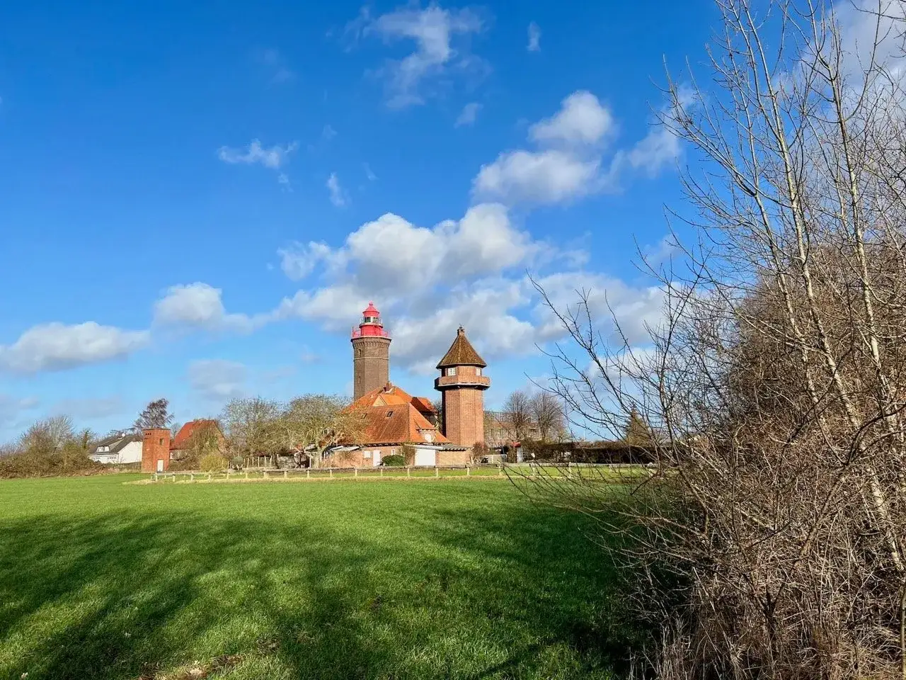 Leuchtturm und Geb&auml;ude mit roten D&auml;chern unter blauem Himmel mit Wolken. Ein gr&uuml;nes Feld und kahle B&auml;ume im Vordergrund. Dahme Geheimtipps f&uuml;r einen Ausflug.