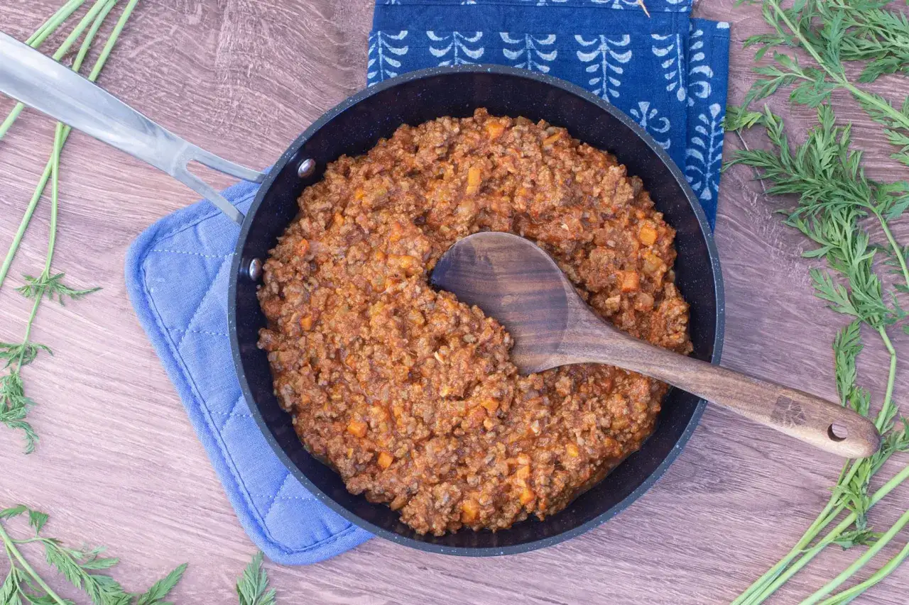 Eine Pfanne mit k&ouml;stlicher, **schneller Bolognese** steht auf einem blauen Topfschutz. Frische Karottengr&uuml;n liegt daneben.